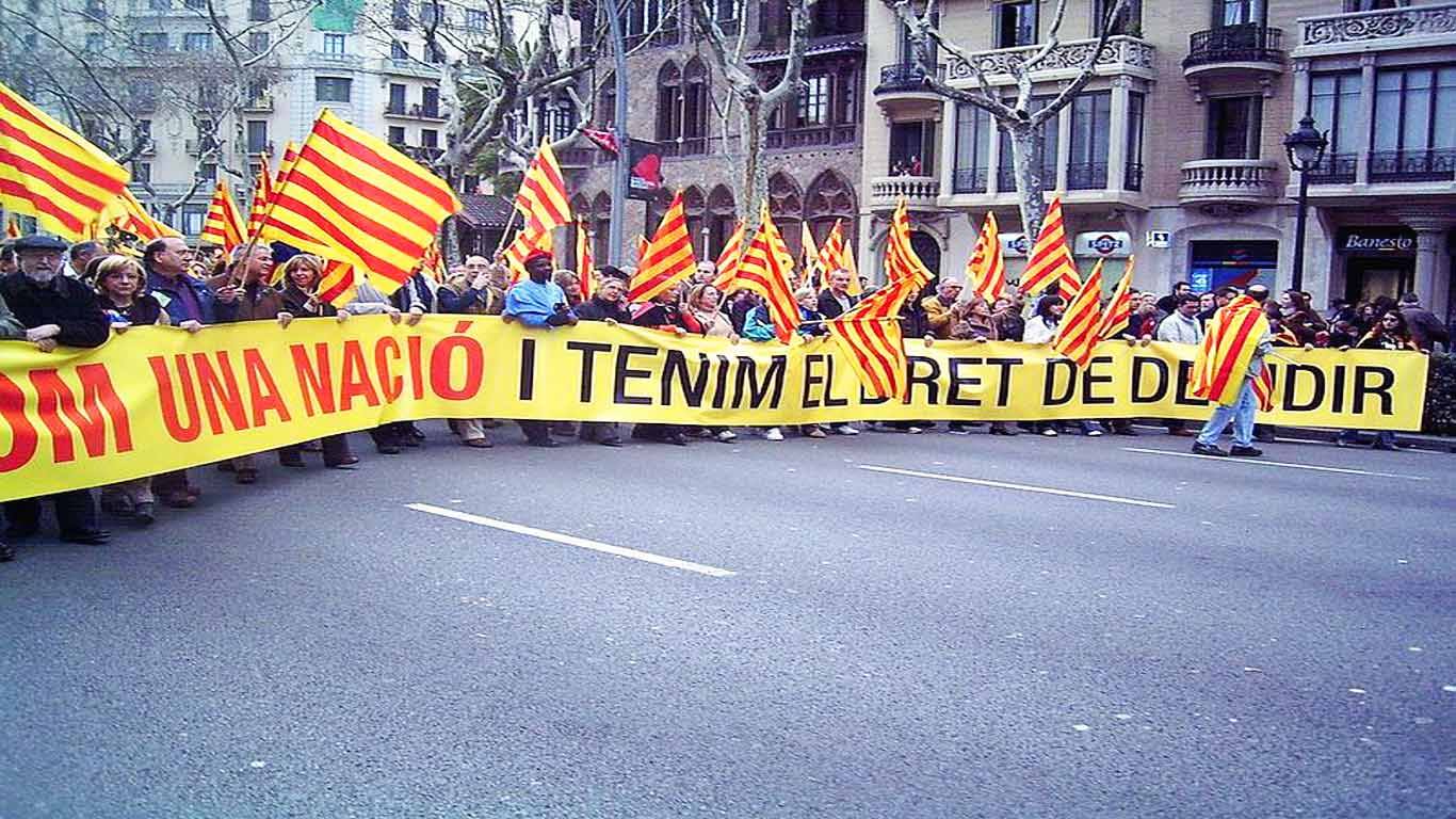 Manifestação em progresso, onde milhares de pessoas erguem bandeiras catalãs e uma tarja de revolta. 