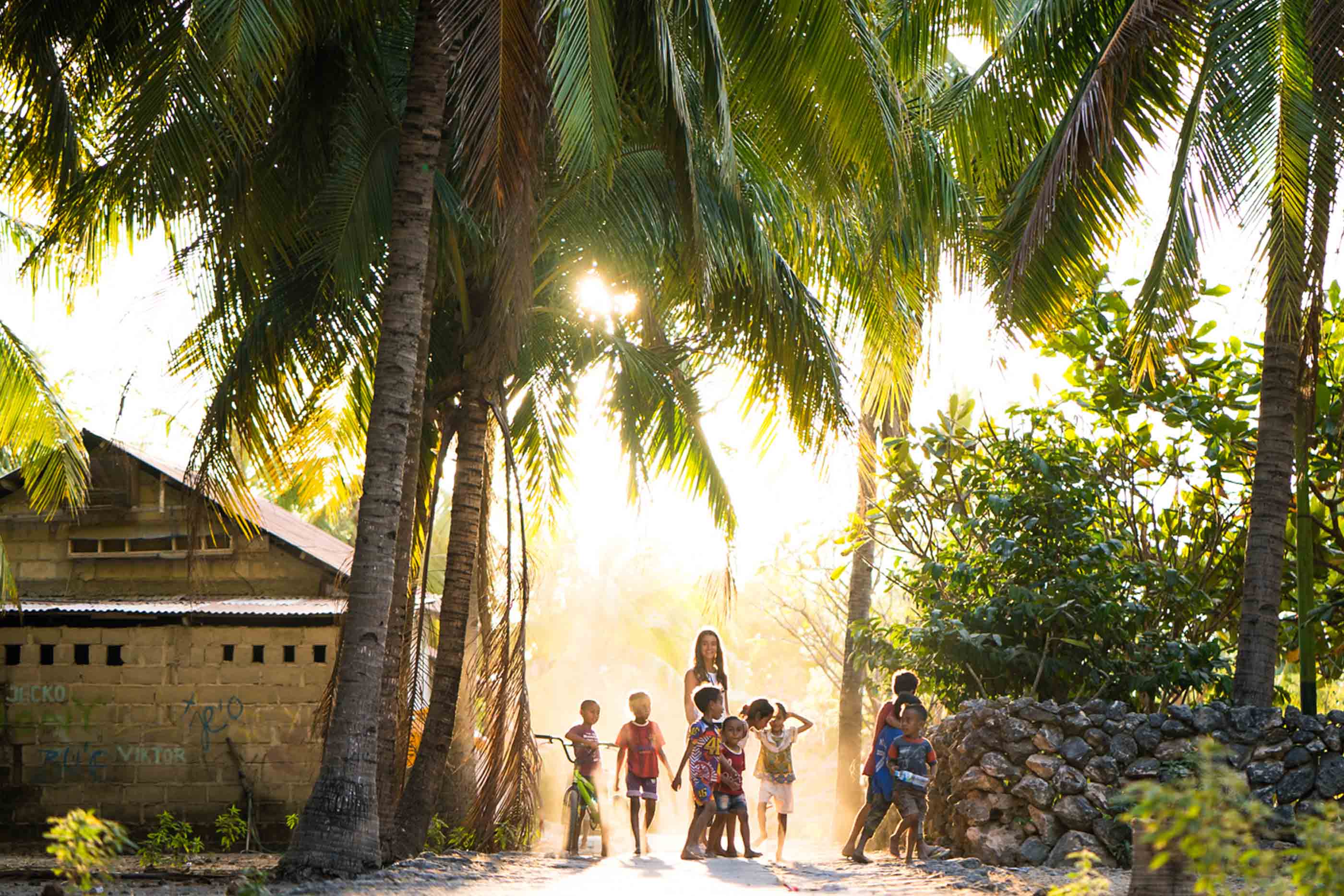 Paisagem tropical com palmeiras à volta e uma casa do loado esquerda da imagem. No centro, crianças e uma senhora.