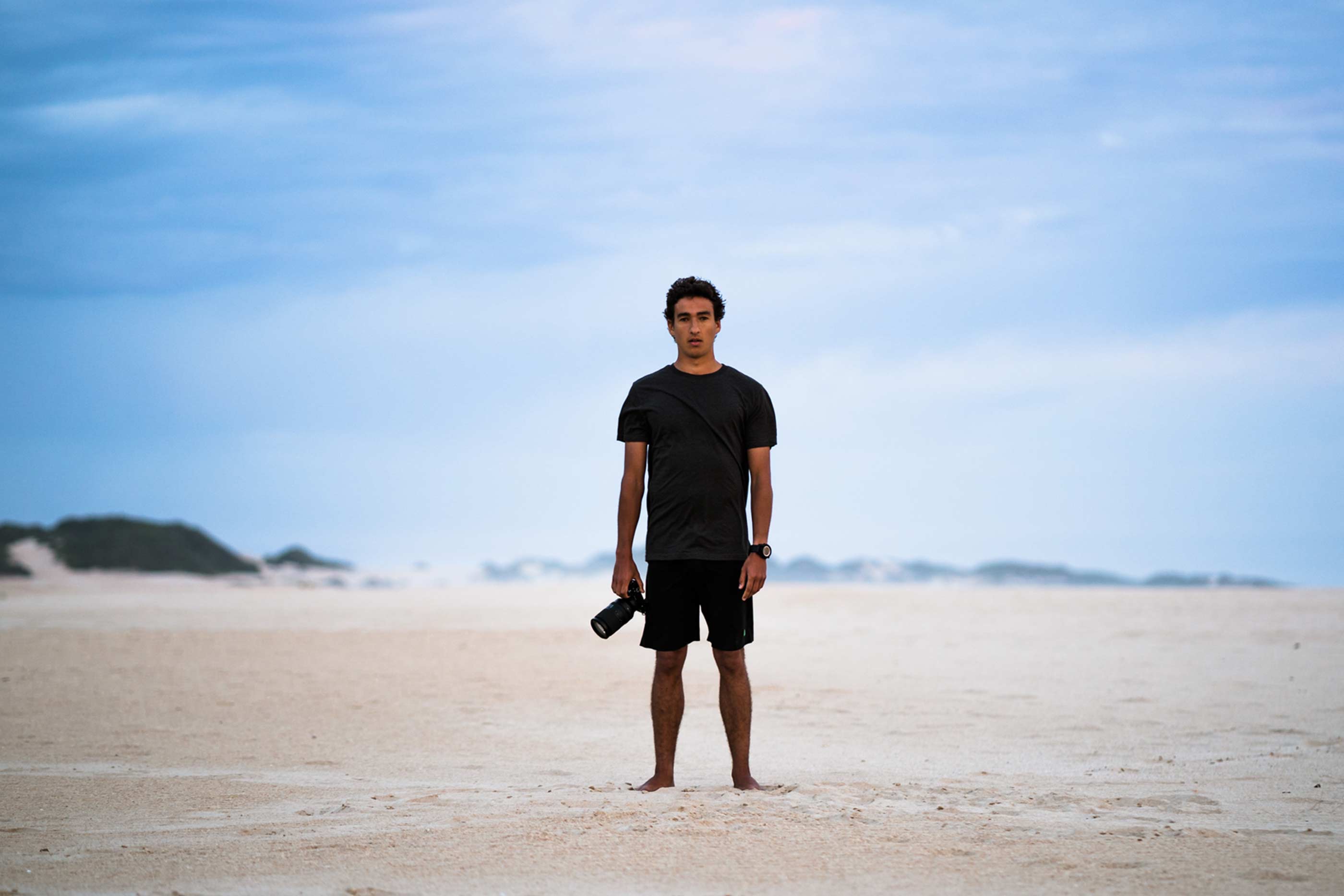 Rapaz, em pé, no centro da imagem, com máquina fotográfica na mão. No fundo, areia e céu azul.