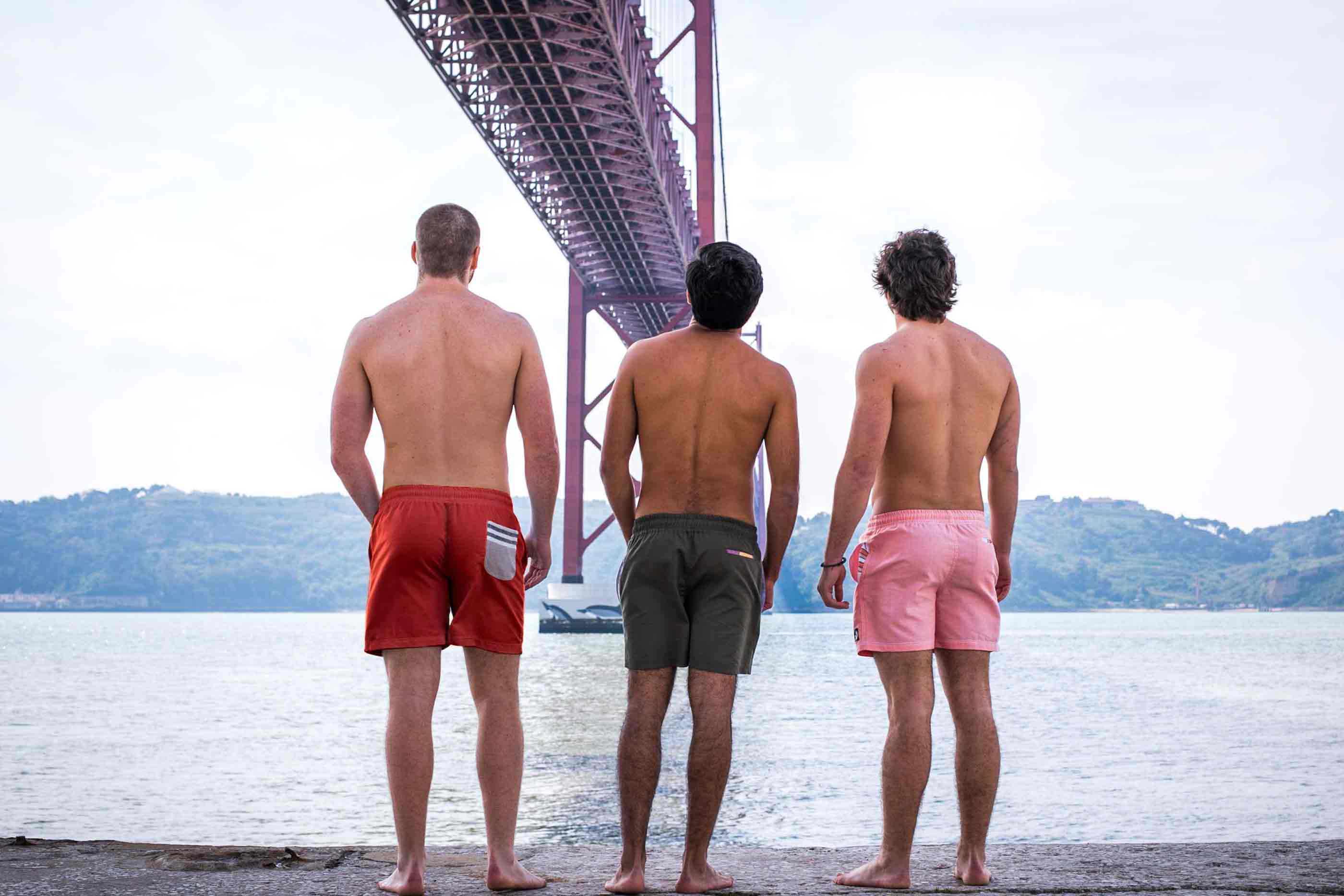 Ao fundo, o mar, colinas e por cima a ponte 25 de abril. Três rapazes de costas com calções de banho de cores diferentes.