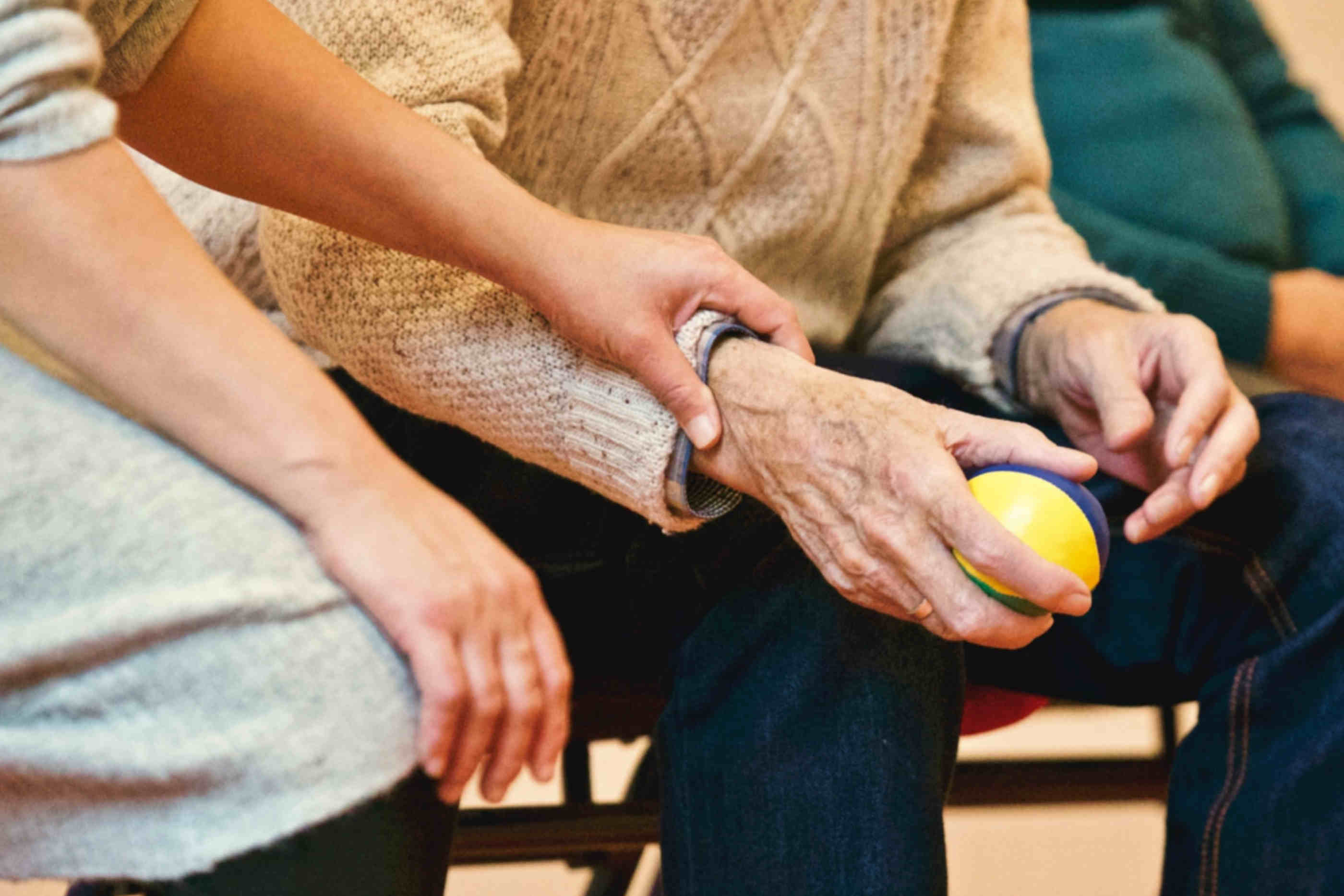Senhora e idoso sentados lado a lado. Senhora mede a pulsação do idoso enquanto este último agarra uma bola anti-stress para as mãos.