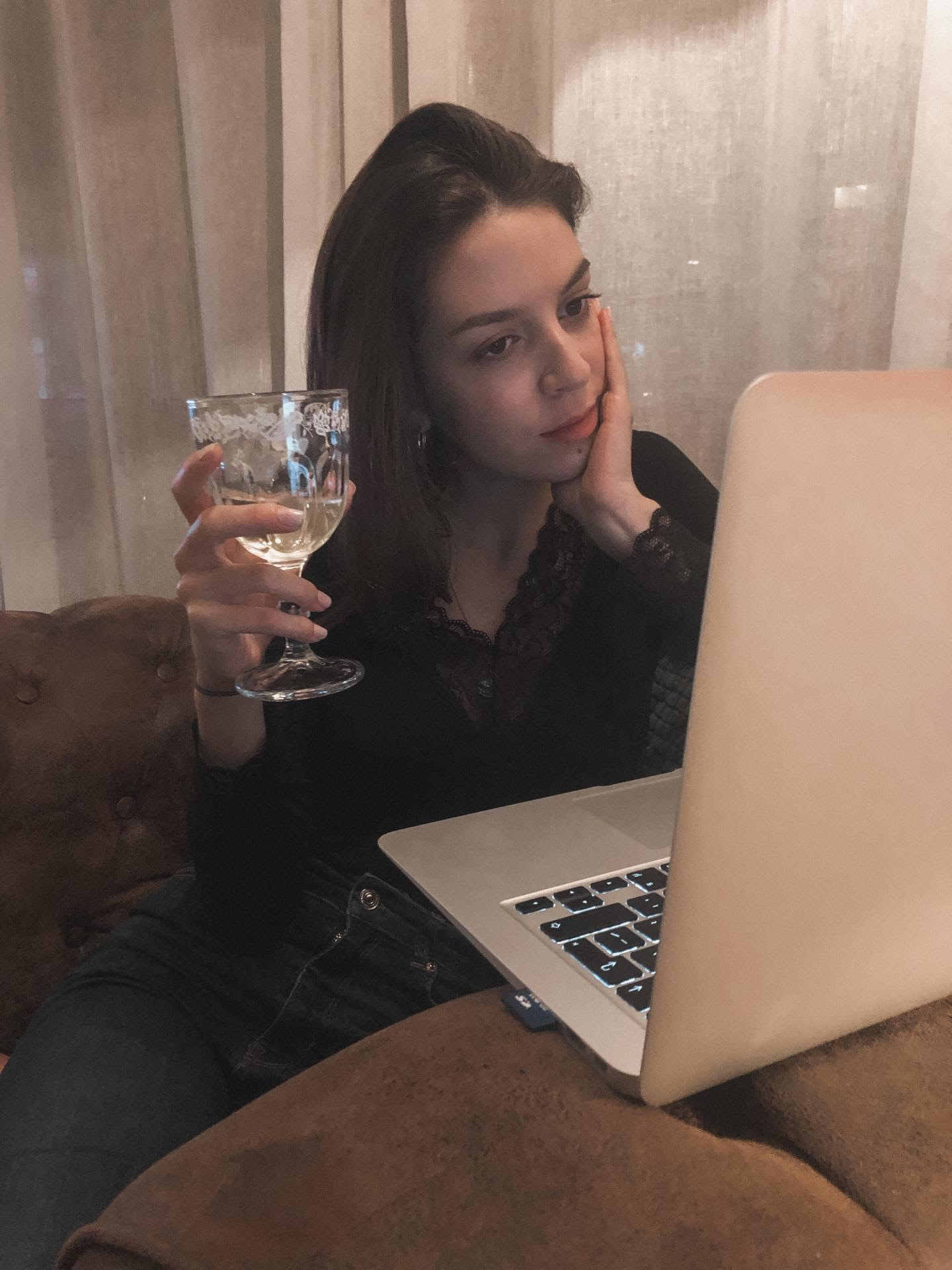 Rapariga sentada no sofá, em frente ao computador, com um copo de vinho branco na mão. 