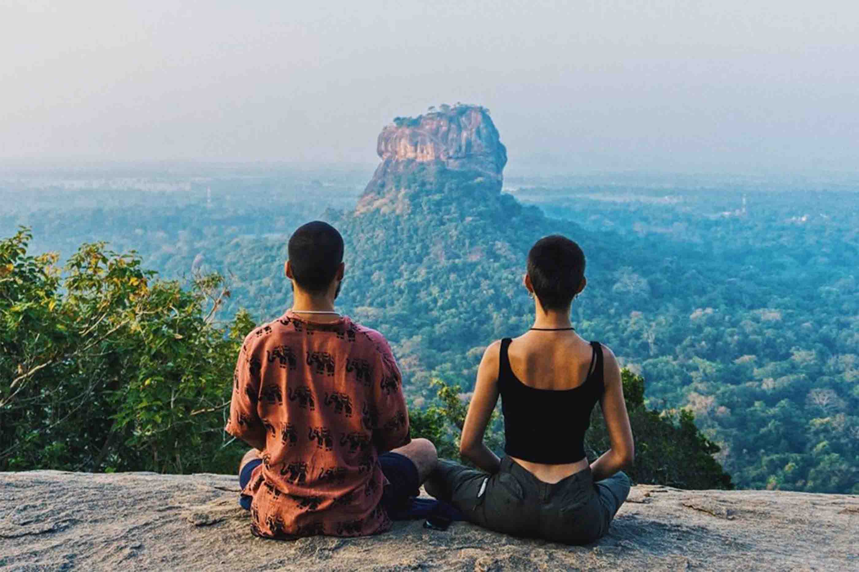 Rapaz e rapariga sentados de costas no topo de montanha com vista verdejante