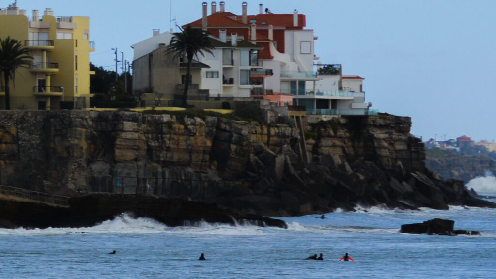Jovens surfistas apanham ondas no mar enquanto ao fundo existe um prédio amarelo e um prédio branco, as ondas batem nas rochas