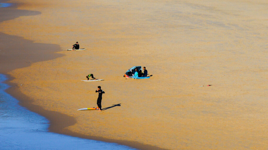 Jovens preparam-se para surfar fazendo um aquecimento no areal em cima das suas pranchas a ver encontra-se um casal debaixo de um guarda – sol azul