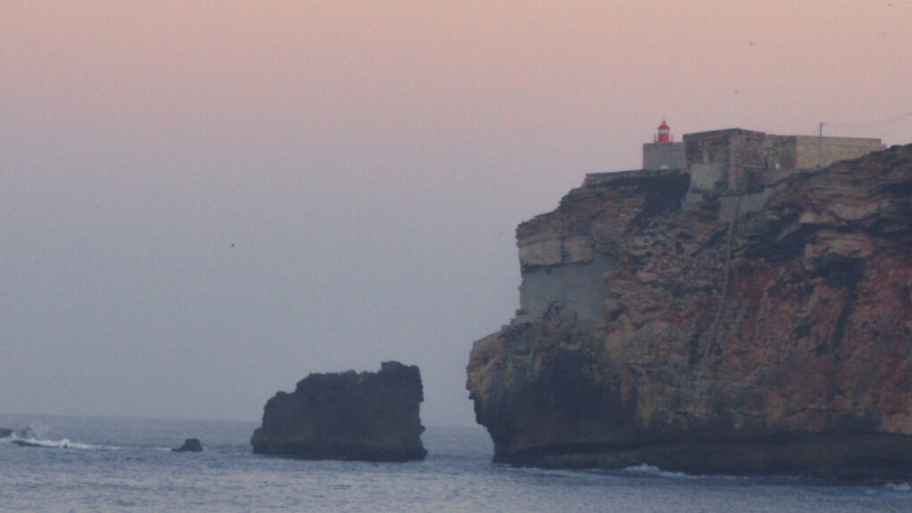 Ao fundo vê-se o mar e as rochas, assim como também se vê um farol