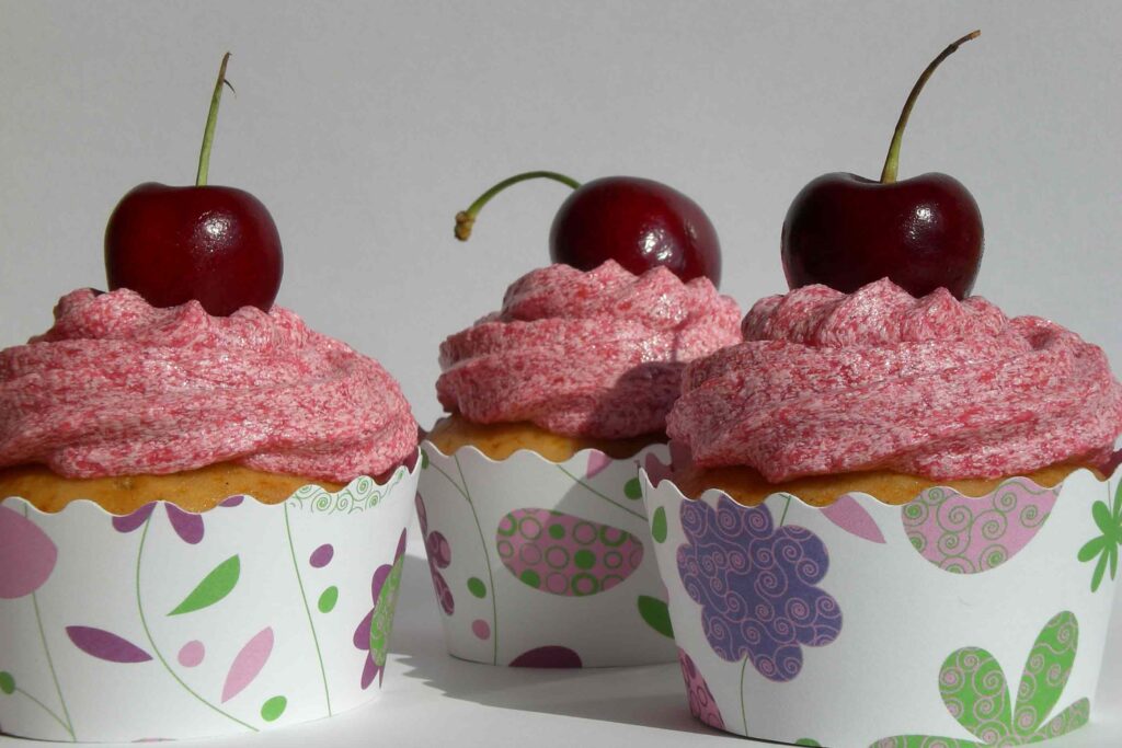 Cupcakes vegan de baunilha e néctar de agave com cobertura de cereja sevidos em formas brancas com flores e formas abstratas rosas, verdes e roxas.