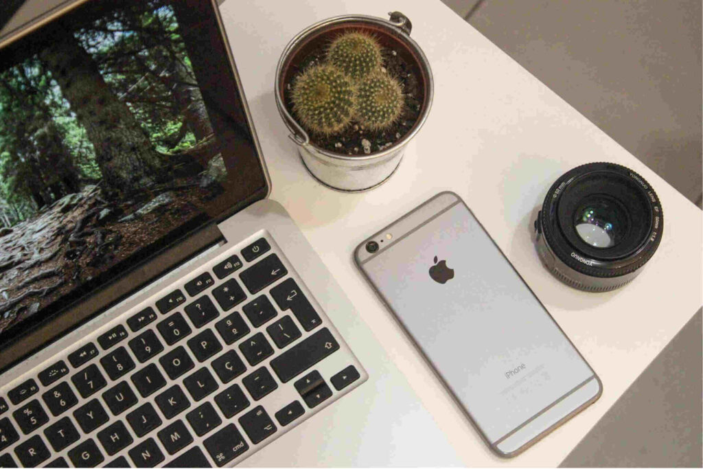 Computador junto de um computador ambos de cor cinzenta em cima de uma mesa com uma lente de camara e um vaso.