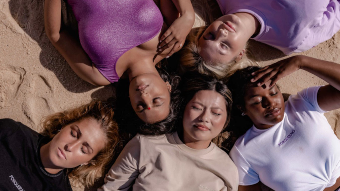 cinco mulheres de diferentes etnias, deitadas na areia em círculo