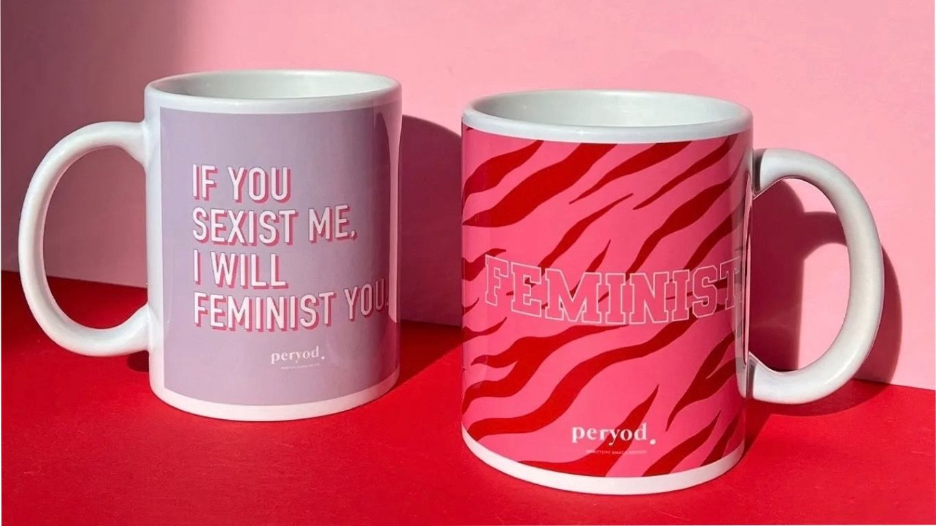 Duas canecas, uma roxa do lado esquerdo que refere “If You Sexist Me, I Will Feminist You” e outra rosa e vermelha do lado direito que refere “Badass. Feminist”
