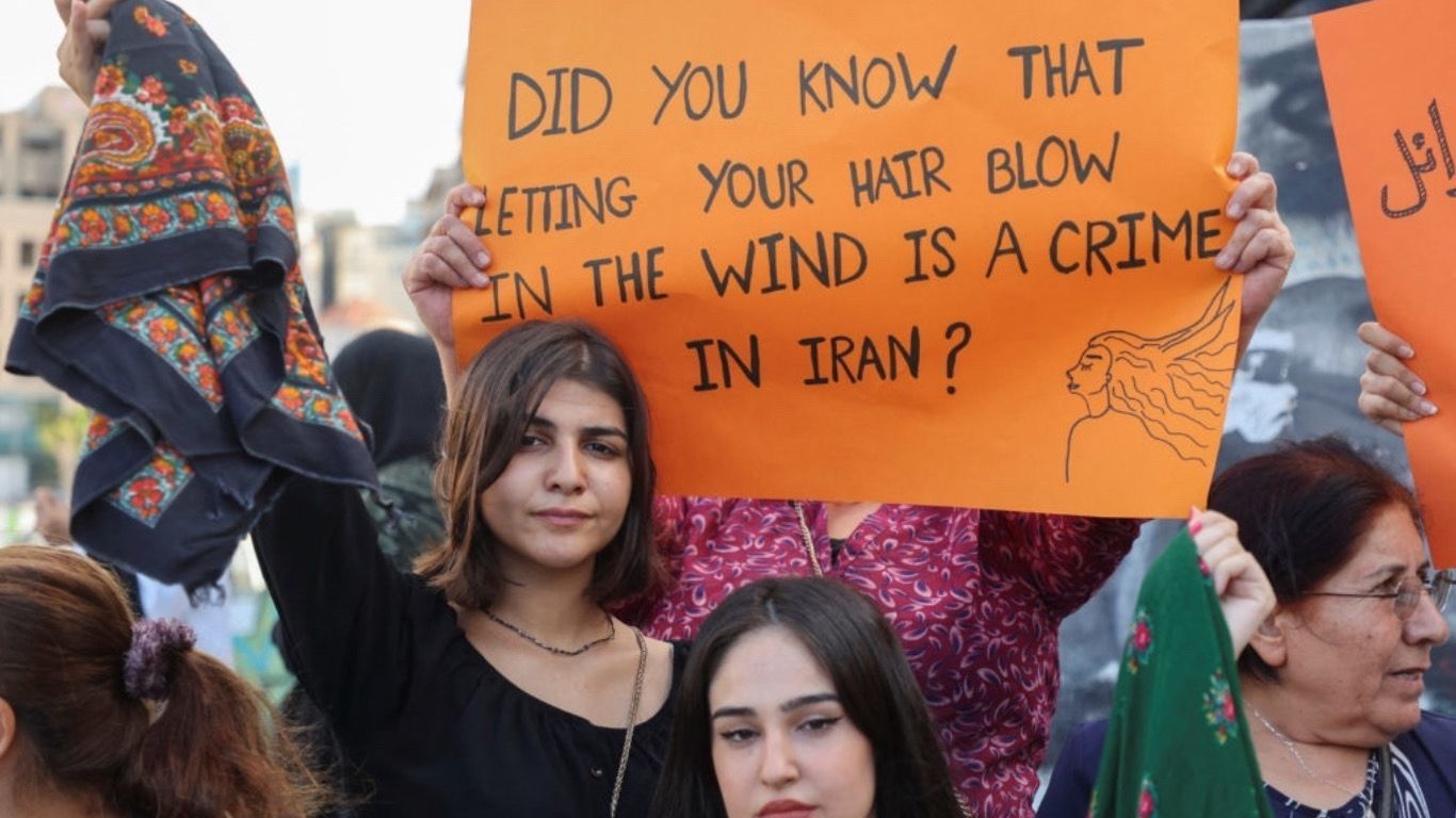 Várias mulheres em manifestação, onde se destacam duas mulheres em primeiro plano com cartaz “Did you know that letting your hair blow in the wind is a crime in Iran?”