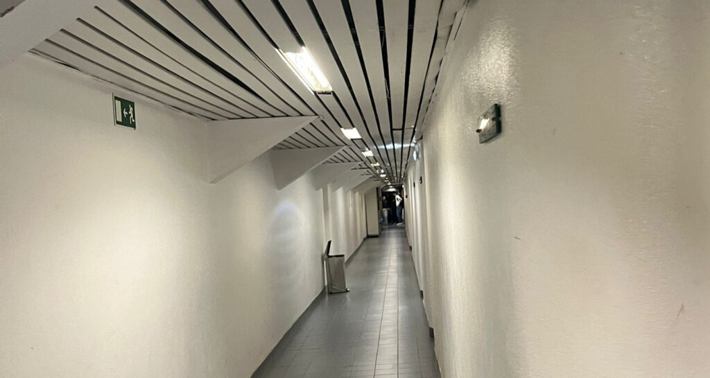 Corredor dos balneários, com paredes brancas e teto com ripas, iluminado por luzes LED.