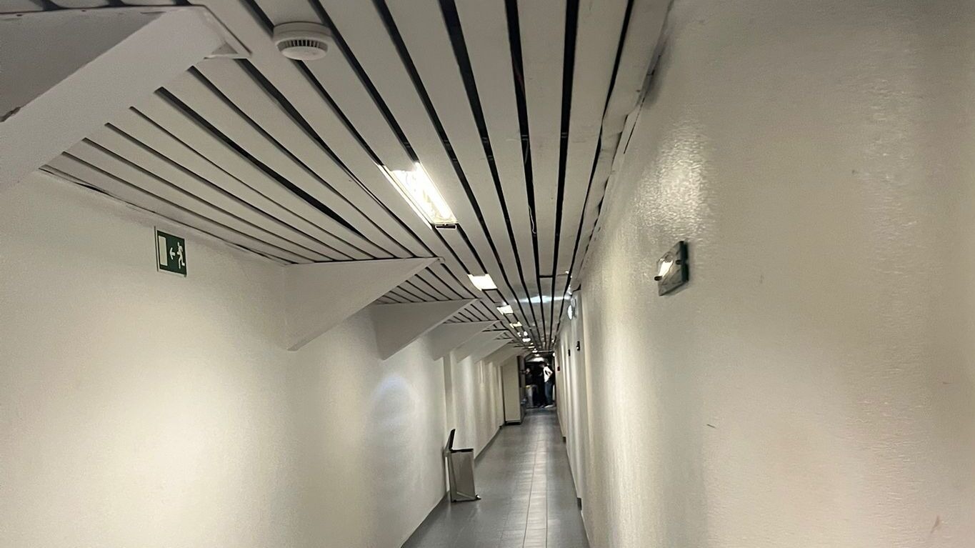 Corredor dos balneários, com paredes brancas e teto com ripas, iluminado por luzes LED.