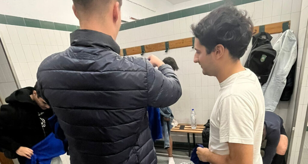 Grupo de jogadores da Católica a prepararem-se num balneário com paredes brancas e bancos de madeira.