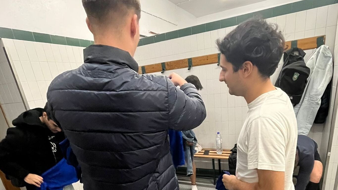 Grupo de jogadores da Católica a prepararem-se num balneário com paredes brancas e bancos de madeira.