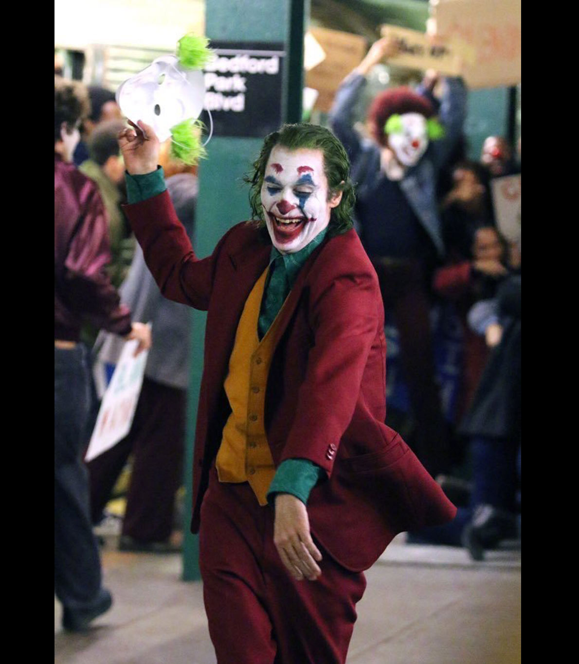 Fotografia do set do filme “Joker”. Nesta fotografia, o ator que representa a personagem Joker, Joaquin Phoenix, aparece já caraterizado, vestido e pintado de palhaço, rindo e segurando uma máscara.