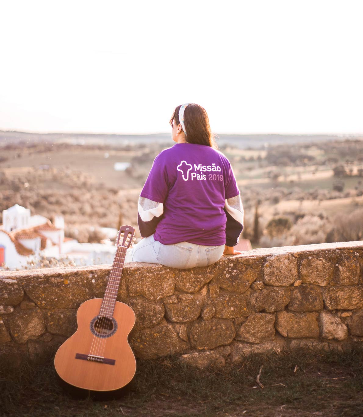 Uma jovem vestida de roxo sentada num muro de pedra a olhar para a vista da cidade e uma guitarra pousada contra o muro.