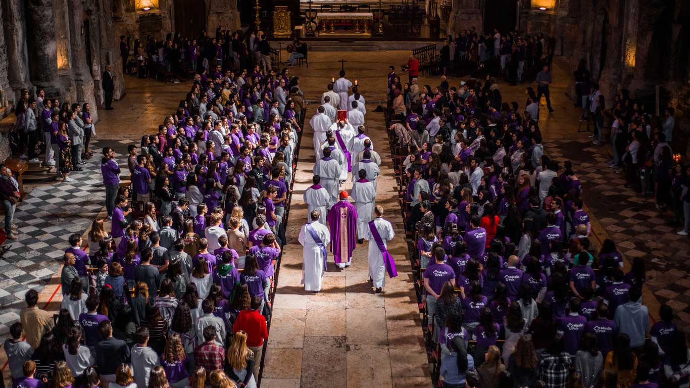 Um grande numero de pessoas a olharem para uma procissão de vários padres vestidos de branco dirigindo-se para o altar