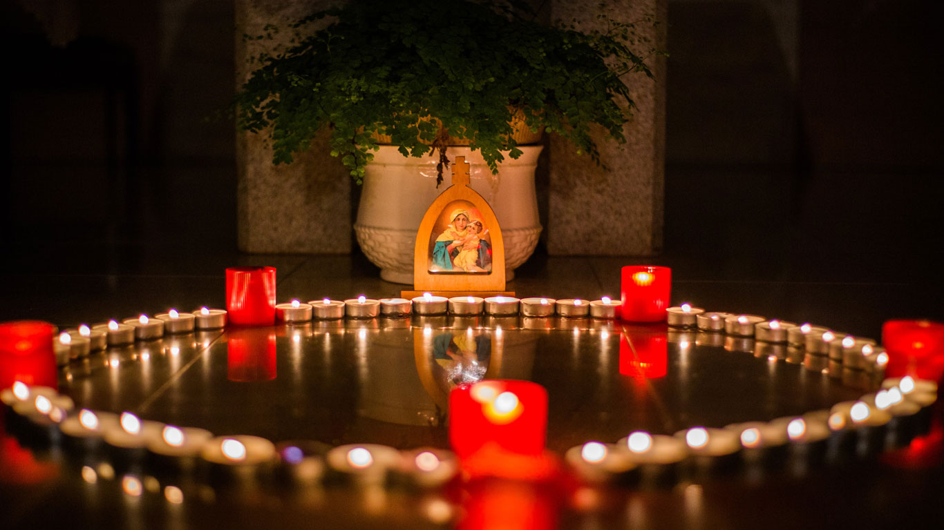 Um circulo de velas acesa com uma imagem de madeira de Nossa Senhora ao fundo pousada à frente de um vaso branco