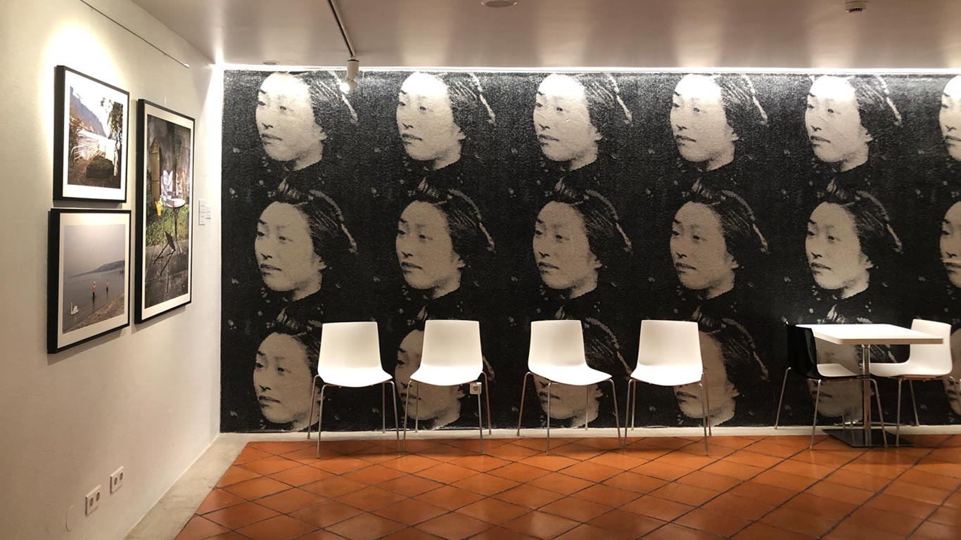 Três paredes, duas delas com fotografias de uma exposição, a terceira, é um moral com pequenos azulejos a formar os rostos de mulheres a preto e branco.