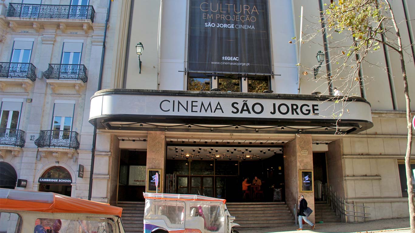 Fachada do Cinema São Jorge, em pleno dia, com dois tuk-tuk parados em frente. Uma pessoa passa, em frente do edifico do cinema, e duas estão sentadas, depois da escadaria do cinema, no café do São Jorge.