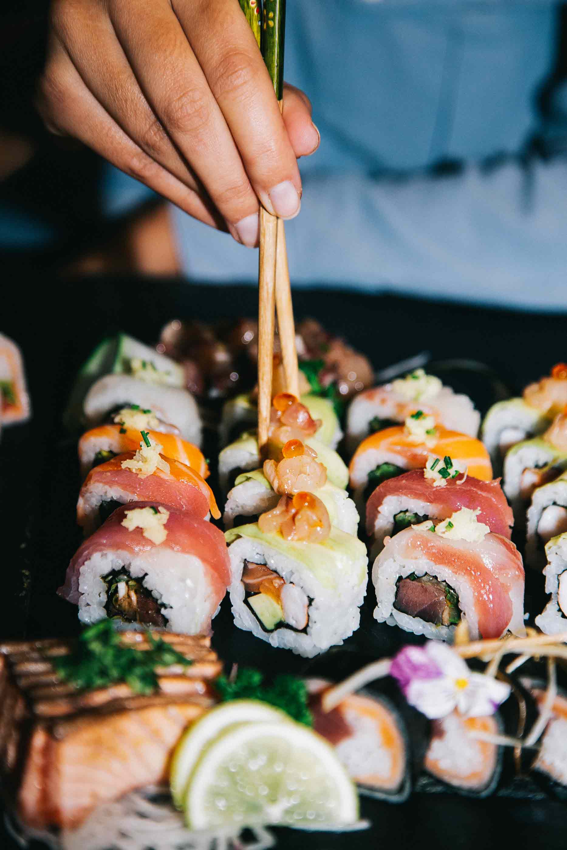 Diferentes peças de sushi sobre um prato preto e o chef Sebastião com os hashi(pauzinhos) a pegar numa peça.