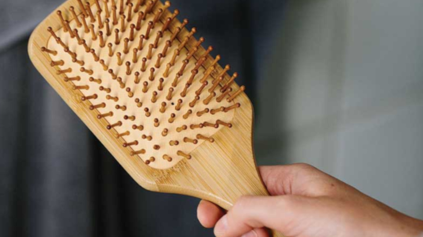 Na imagem observa-se uma mão fechada a agarrar numa escova de cabelo de bambu.