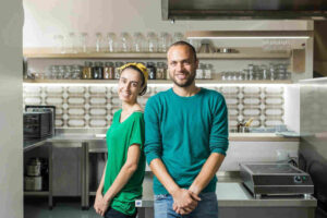 Rapariga e rapaz sorriem na cozinha do seu restaurante, com camisolas verdes que simbolizam o conceito do seu restaurante 100% sustentável.