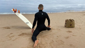 Surfista a alongar com a prancha de surf ao lado, utilizando um saco de cartão reutilizável