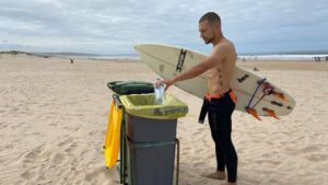Surfista a reciclar uma garrafa de plástico na praia, enquanto segura a prancha de surf