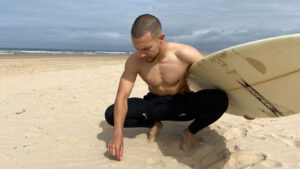 Surfista agachado a apanhar uma beata de cigarro da areia, segurando uma prancha de surf