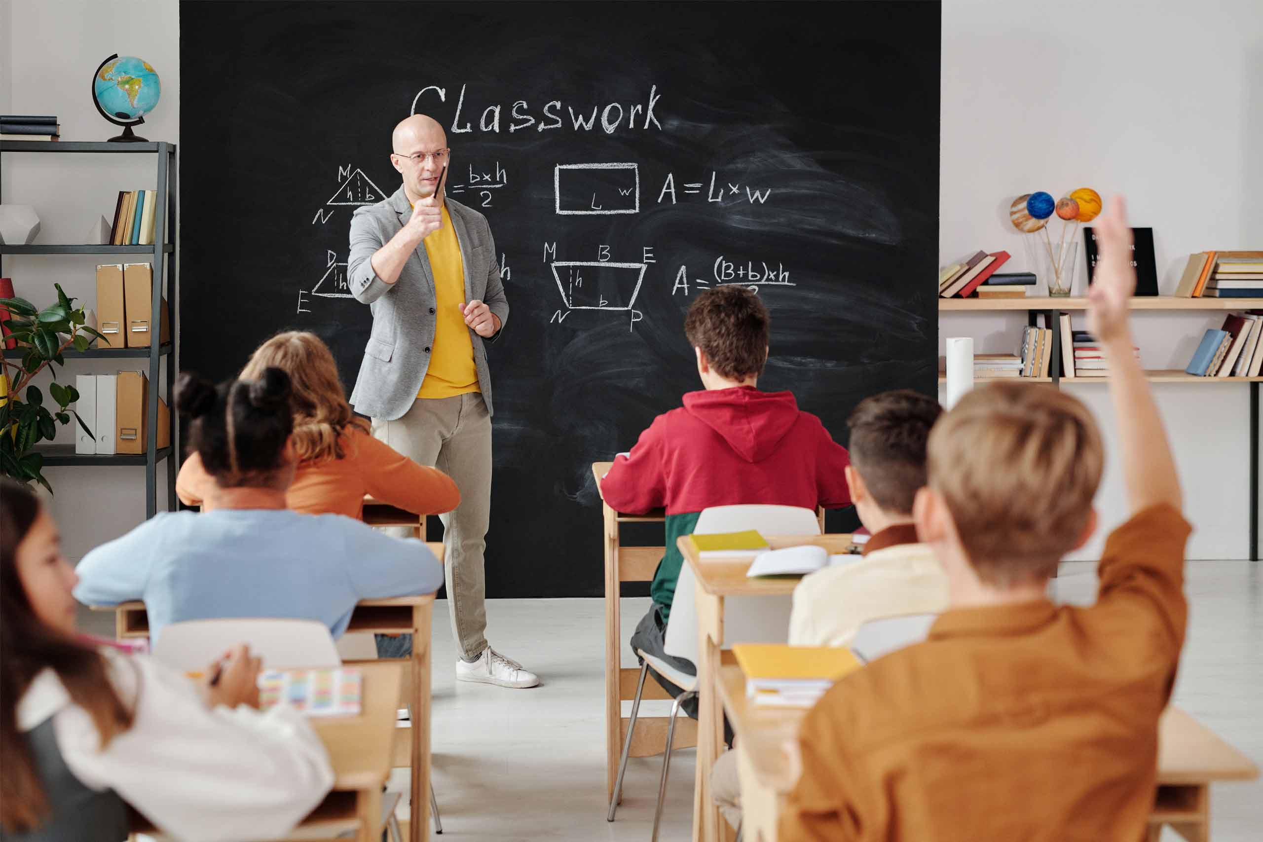 Professor em sala de aula indica a um aluno com a mão levantada para responder a uma questão colocada
