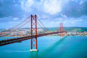 Ponte 25 de Abril em Lisboa vista de perfil, estrutura vermelha sobre o rio Tejo num dia azul com nuvens