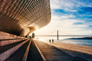 Fachada ondulante do MAAT ao nascer do sol, com duas pessoas a correr junto ao Tejo e a ponte 25 de Abril ao fundo.